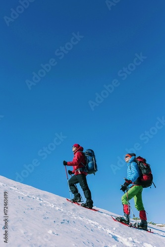 Climber with backpack trekking poles snowshoes rises to the top of the mountain in the snow on a background of beautiful blue sky. 