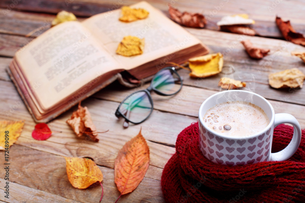 Cup hot cappuccino coffee on wooden table with autumn leaves ,book and eyeglasses.Autumn mood concept.Warm autumn picture .Selective focus