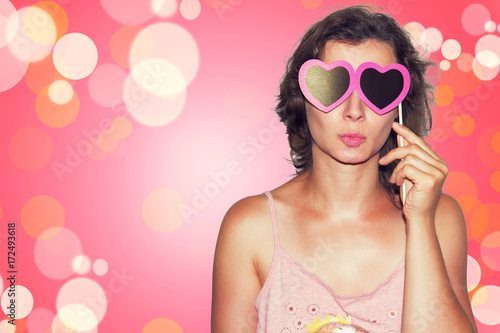 Beauty Young fashion model Girl with Valentine heart paper glasses on pink background. Love. Valentines Day gift.