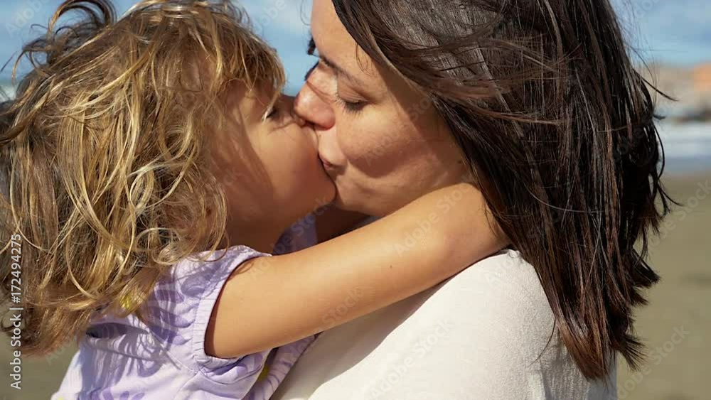 Woman kissing little blond daughter smiling in front of the ocean looking camera slow motion Woman kissing little blond daughter smiling in front of the ocean looking camera slow motion