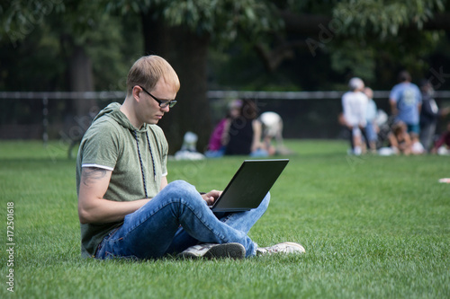 Young man in glasses working on laptop sitting on the grass in park. Maybe student or hacker © evgenzz