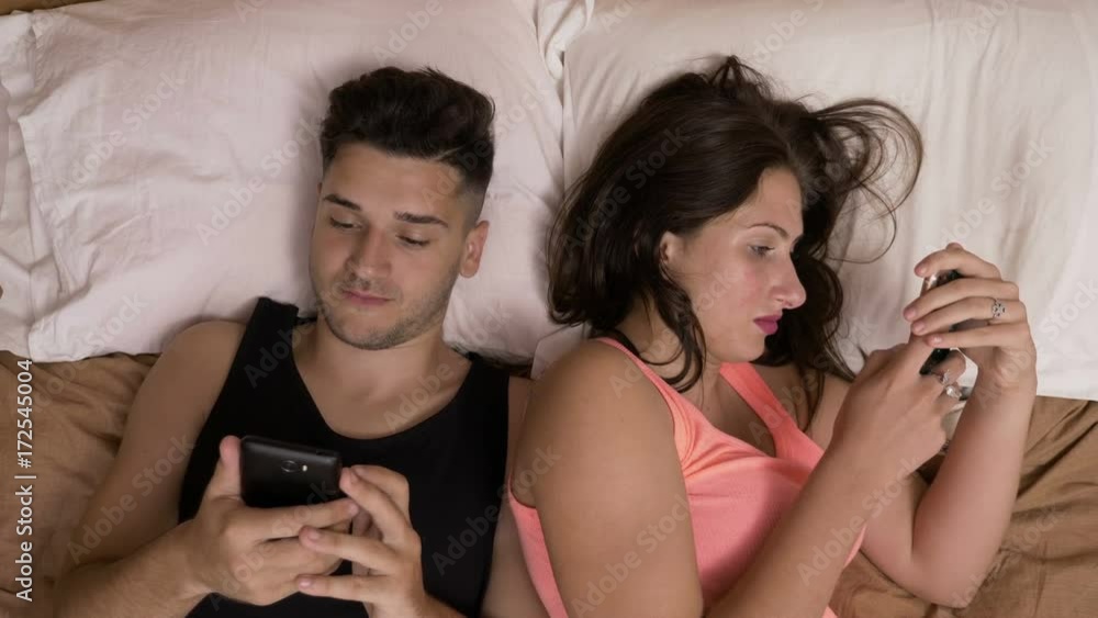 Young couple laying in bed chatting on social media addicted to smartphones ignoring each other Young couple laying in bed chatting on social media addicted to smartphones ignoring each other