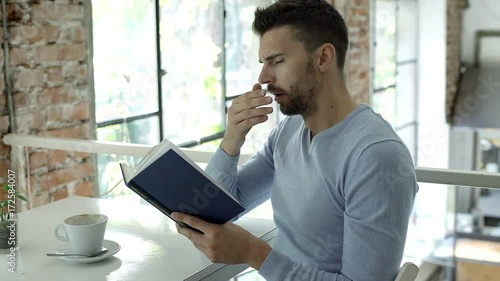 Handsome man yawning while reading book in the cafe, steadycam shot
