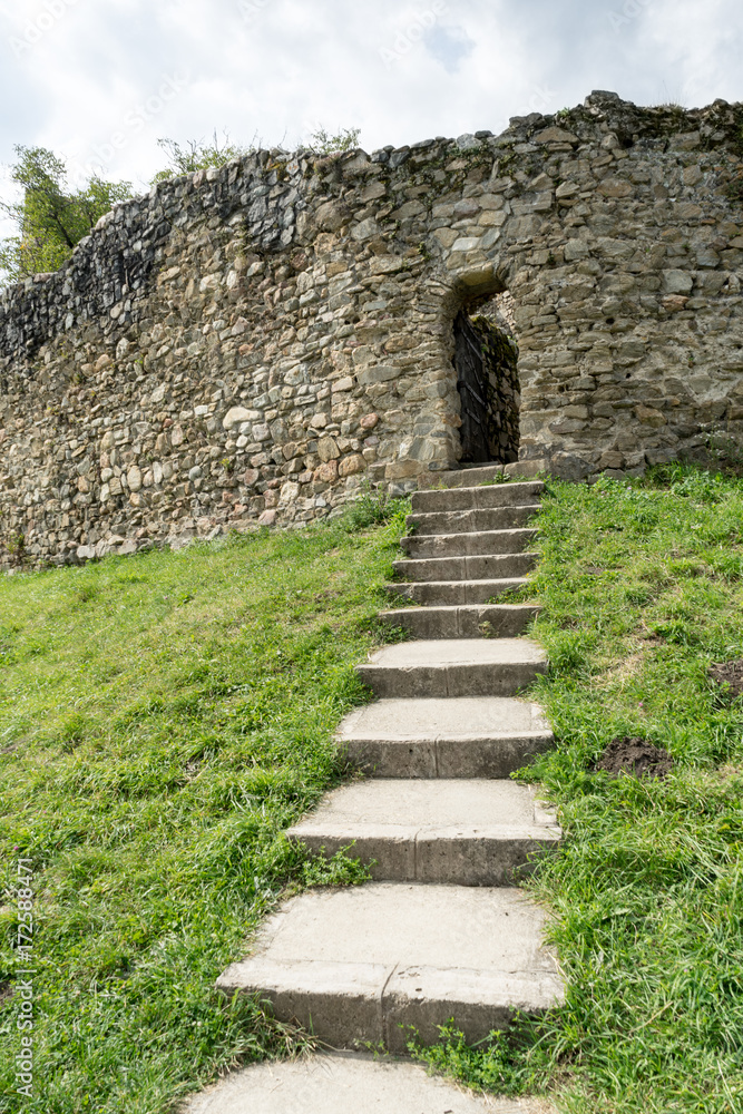 Old stone arched door of ruined ancient castle at Cisnadioara fortress in Sibiu, Romania. Stone wall fortress with stairs leading to door. Green grass all around.