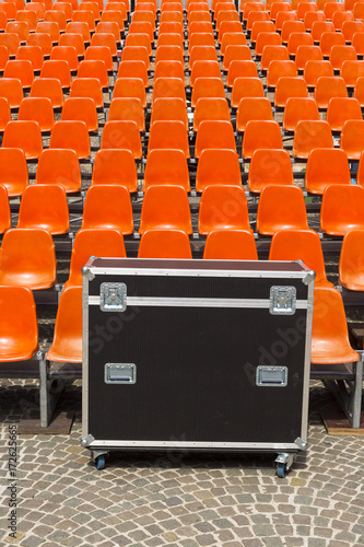 Musical instrument black box on sett ground in front of vacant red plastic seat rows. Red plastic seats and closed concert equipment black box