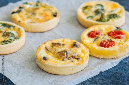Set of savory mini tarts on a baking paper. Vegetable quiches with tomatoes, mushrooms, herbs, broccoly. Blue stone background. Close up and copy space.