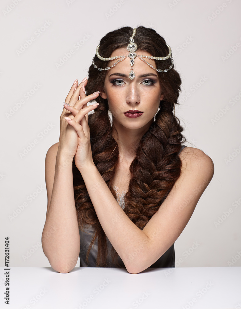 Fototapeta premium Beauty portrait of young woman in pearl diadem. Brunette girl with braids hairdo and day female makeup sit at white table