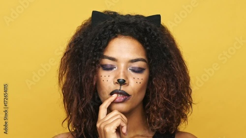 Tricky african lady with cat make-up and ears smiling and flirting isolated over yellow photo