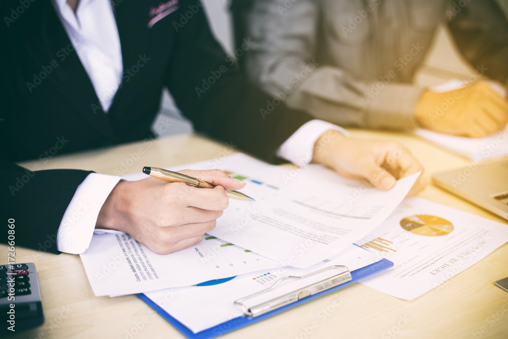 Asian businesswomen holding a pen and analysis documents on office table with laptop computer and graph financial diagram working in the background Asian businesswomen holding a pen and analysis documents on office table with laptop computer and graph financial diagram working in the background