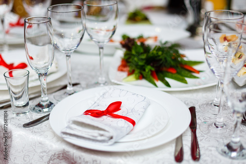 Rolled table napkin tied with red ribbon on white empty plate on table. Table setting, free space. Table served for wedding banquet, close up view