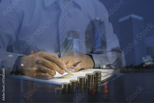 Double exposure businessman writing with stacked of money coins and night ciyt on wood table photo