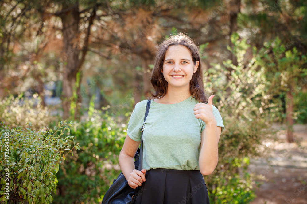 Pretty teenage girl looking at camera, smiling and showing thumbs up outdoors. Portrait of a beautiful girl. Pretty teenage girl looking at camera, smiling and showing thumbs up outdoors. Portrait of a beautiful girl.
