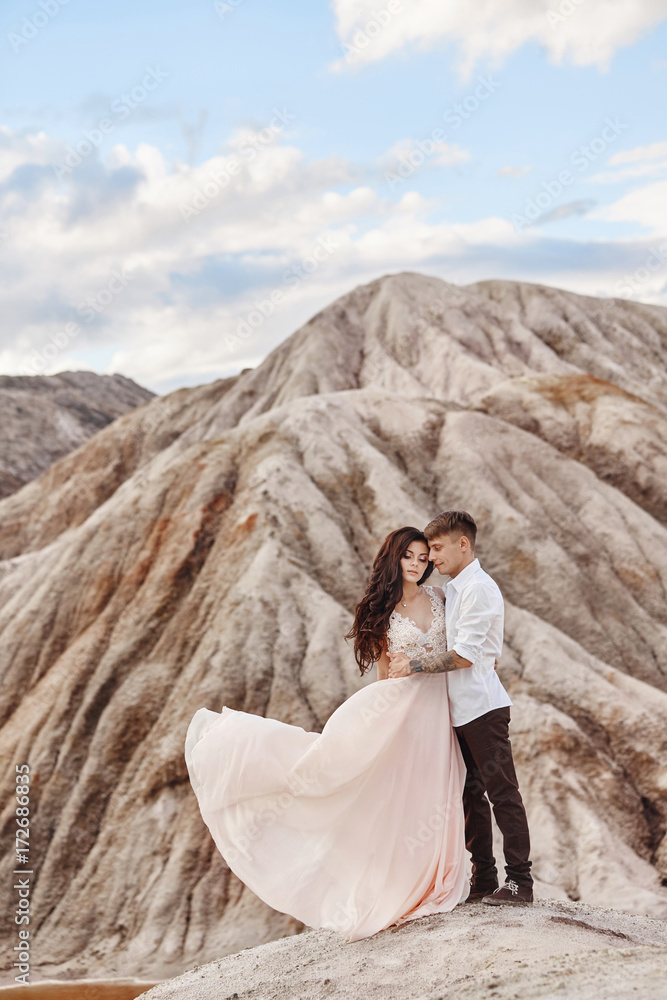 Couple stands on the precipice of the mountain and the red lake and hugging. The bride and groom in love, the woman in long dress in the arms of men