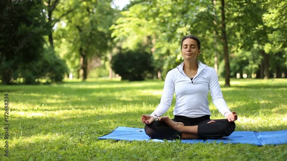 Full of joy young women wearing sports clothes blue shirt and black tights in lotus position meditating in the quiet scenery of late summer in the park Full of joy young women wearing sports clothes blue shirt and black tights in lotus position meditating in the quiet scenery of late summer in the park