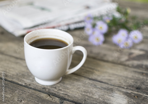 White cup of coffee and newspaper and flowers on old  wooden table. High angle view and selective focus