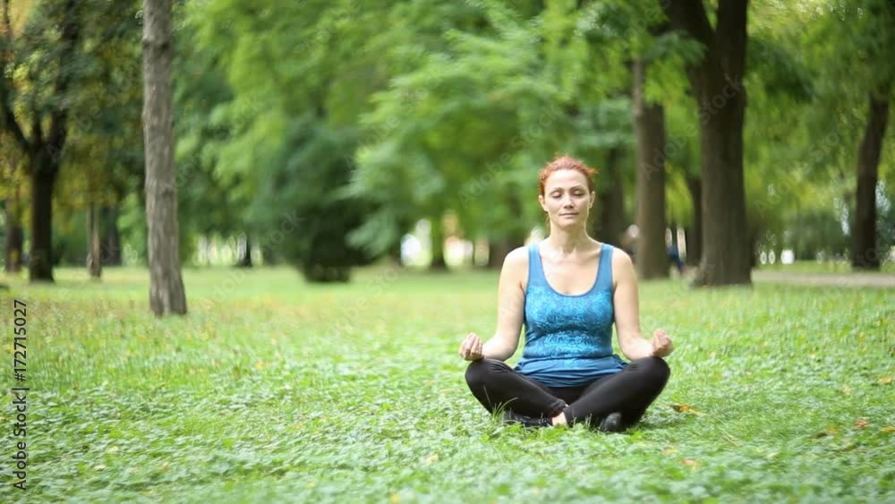 Full of joy young women wearing sports clothes blue shirt and black tights in lotus position meditating in the quiet scenery of late summer in the park, camera is gently moving / Joy of doing yoga Full of joy young women wearing sports clothes blue shirt and black tights in lotus position meditating in the quiet scenery of late summer in the park, camera is gently moving / Joy of doing yoga