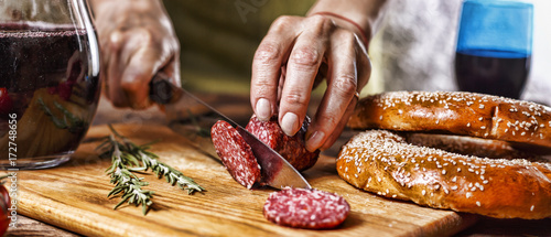 Traditional Italian red wine, salami, rosemary, bread. Close up of a person's hand cut salami on a kitchen board.