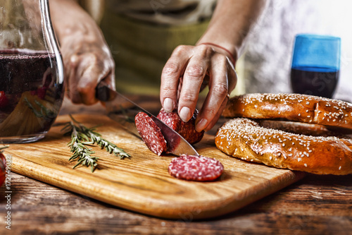 Traditional Italian red wine, salami, rosemary, bread. Close up of a person's hand cut salami on a kitchen board.