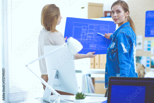 Two young woman standing near desk with instruments, plan and laptop.