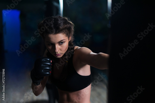 Young fighter boxer fit girl wearing boxing gloves in training with heavy punching bag in gym. Moment before punch. Woman power © Igor Kardasov