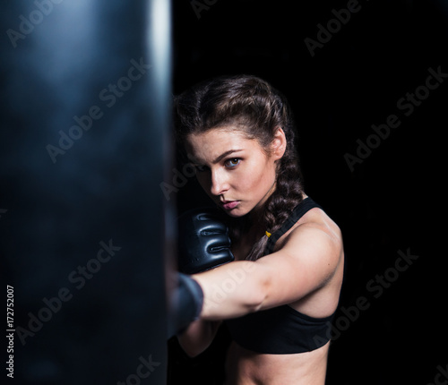 Young fighter boxer fit girl wearing boxing gloves in training with heavy punching bag in gym. Isolated on black background
