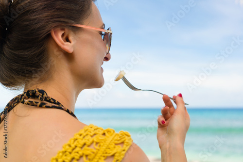 Woman on tropical beach eating banana with a fork looking at the sea