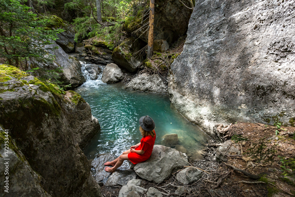 Une jeune fille hipster avec une robe rouge et un chapeau assise au bord d'une piscine naturelle verte au milieu des bois Une jeune fille hipster avec une robe rouge et un chapeau assise au bord d'une piscine naturelle verte au milieu des bois