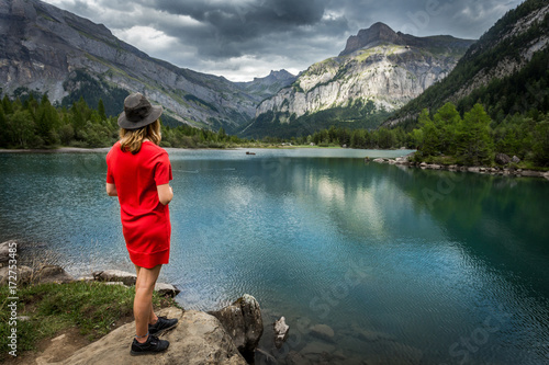 Une jeune femme blonde de dos avec une robe rouge et un chapeau au bord d'un lac de montagne