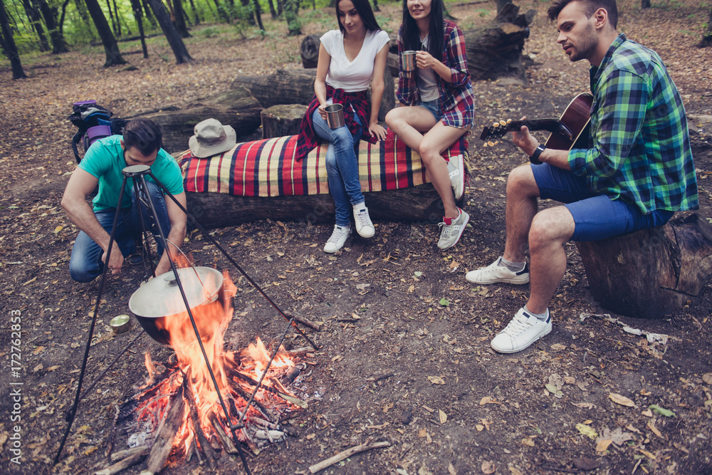 Fire place, four serene tourists are sitting around, girls drink tea, guys play guitar and open the canned goods, spring campsite Fire place, four serene tourists are sitting around, girls drink tea, guys play guitar and open the canned goods, spring campsite