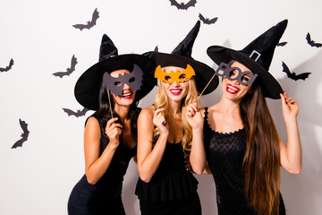 Group of three diverse charming coquettes in dark masquerade elegant dresses, masks on eyes, smiling, enjoying near decorated wall with bats, toothy beaming smiles, scary bright cosmetics hot figures
