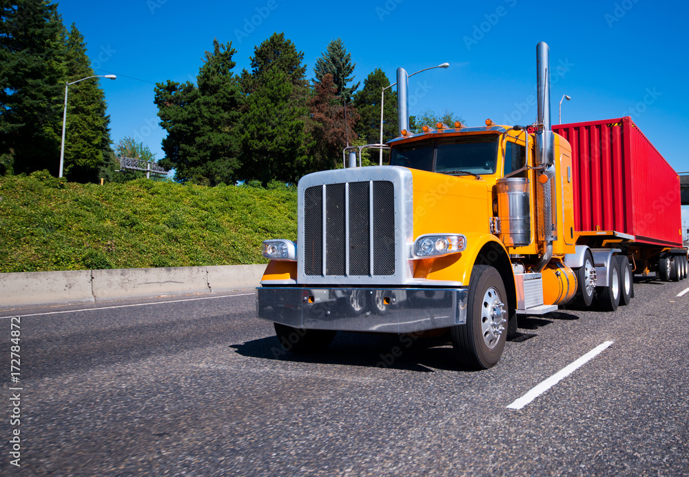 Classic orange big rig semi truck with red container on flat bed trailer running on the road