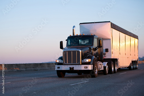 Obraz na plátně Dark big rig semi truck with bulk trailer running on highway in sunset light