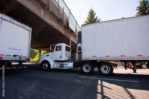 Day cab big rig semi truck going with reefer trailer with refrigeration unit under the bridge on roadside with traffic