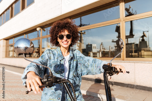 Image of happy curly woman in sunglasses sitting on motorbike