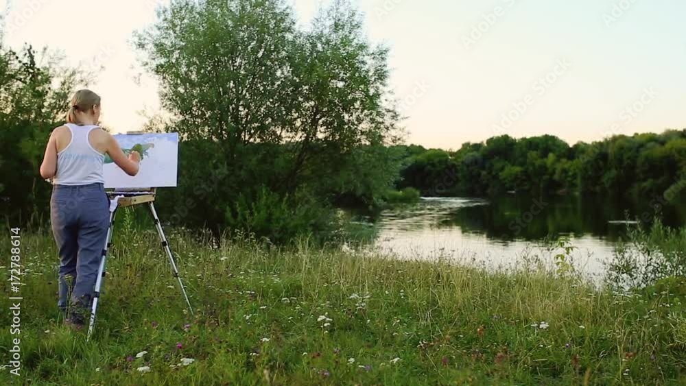 A woman artist with a brush and polytray in her hands draws on canvas against the backdrop of nature. The artist draws on the easel. Artist at work A woman artist with a brush and polytray in her hands draws on canvas against the backdrop of nature. The artist draws on the easel. Artist at work