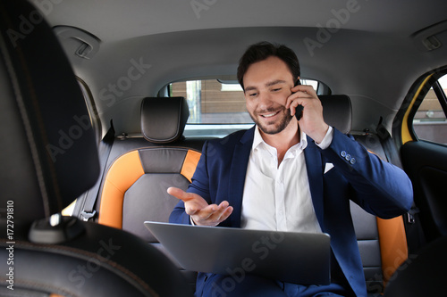 Handsome man with laptop talking on phone while sitting in taxi car © Africa Studio
