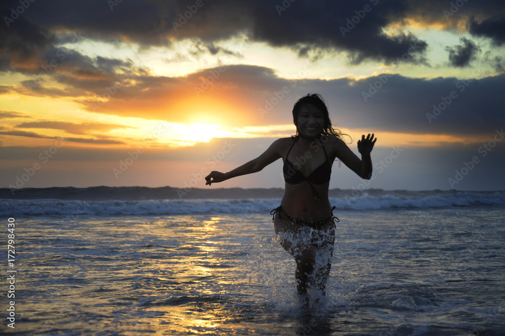 Silhouette of young happy sexy Asian woman in bikini running excited on sunset beach