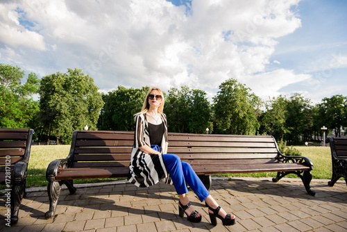 Красивая, стильная девушка со светлыми волосами сидит на скамейке в парке в черной майке и синих брюках 