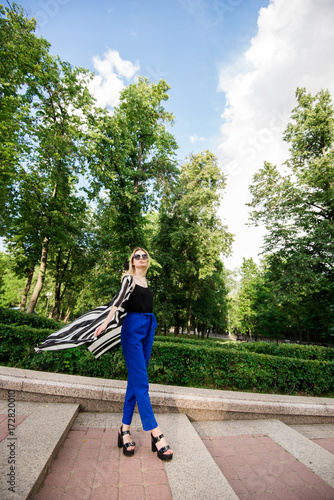 Красивая, стильная девушка стоит в парке в черной майке и синих брюках. Девушка со светлыми волосами в солнечных очках 