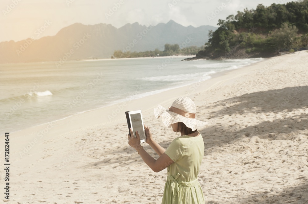 Young asian woman standing on the beautiful nice blue beach with white sand and taking photo with tablet.