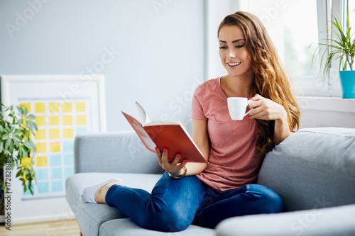 Fotografija Cheerful attractive young woman reading a book and drinking coffee, sitting on s