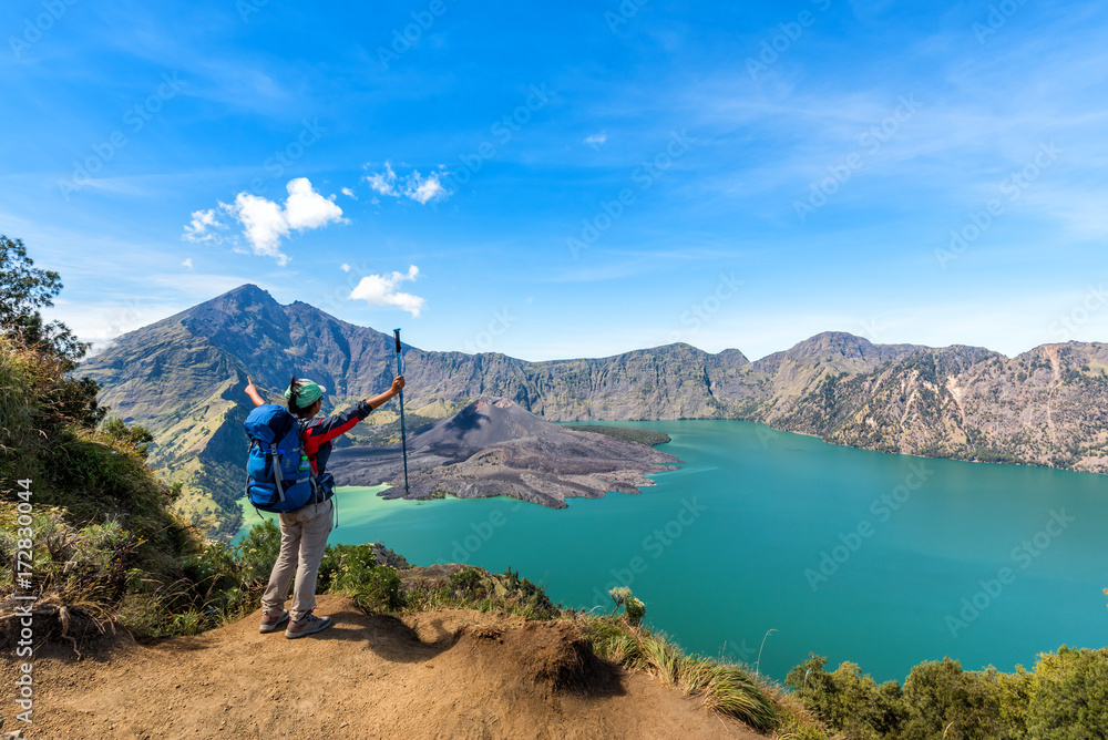 Hiker woman with blue backpack and trekking pole standing and enjoy with volcano Baru Jari Lake Segara Anak and Mount Rinjani summit view after finished climbing at Rinjani mountain. Lombok, Indonesia