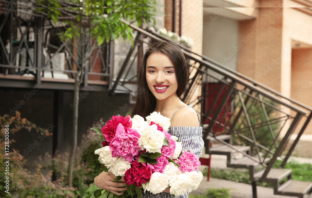 Fototapeta premium Young cheerful woman with beautiful bouquet of peonies standing on street