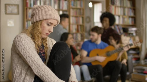 Young woman in shared apartment feeling sad and isolated while others socialize in background.