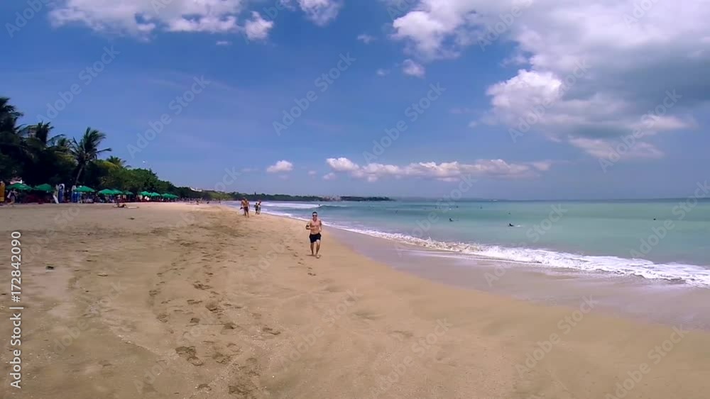 Young athletic man jogging along the shore and waving on beautiful tropical beach. Panoramic scenic view ta the ocean coastline. Travel concept Young athletic man jogging along the shore and waving on beautiful tropical beach. Panoramic scenic view ta the ocean coastline. Travel concept