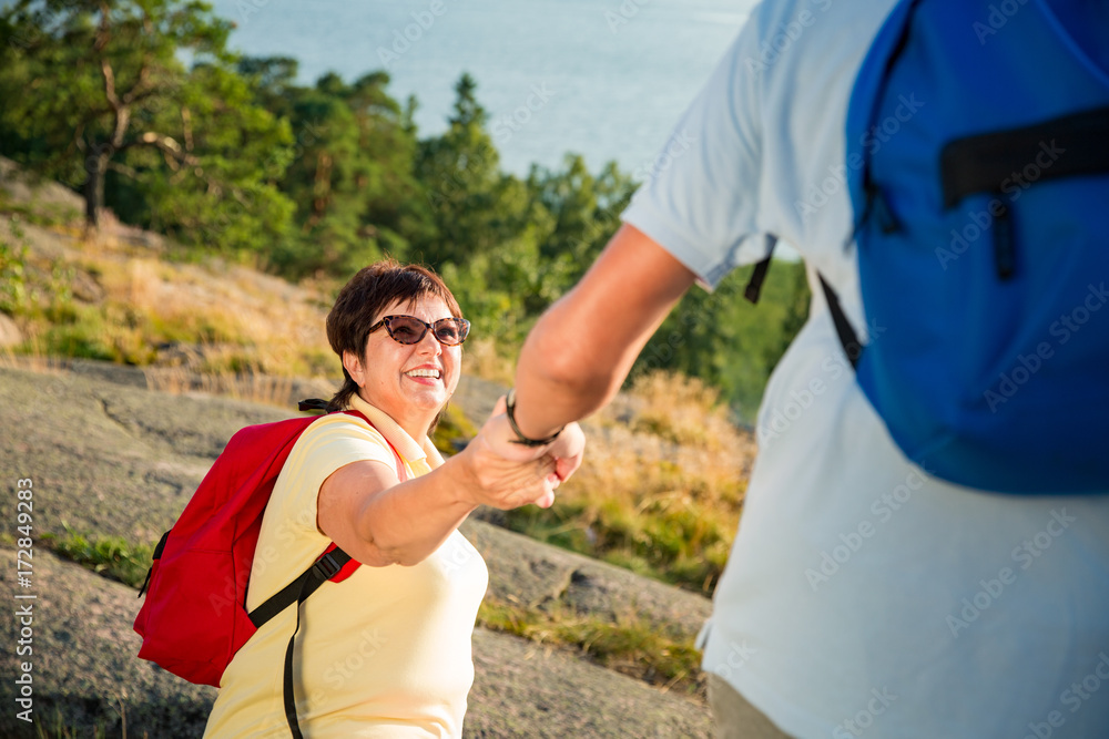 Active senior couple hiking on the top of rock. Mature man helping woman climbing up. Happily smiling. Scenic view of gulf and sea. Healthy lifestyle. Finland. Active senior couple hiking on the top of rock. Mature man helping woman climbing up. Happily smiling. Scenic view of gulf and sea. Healthy lifestyle. Finland.