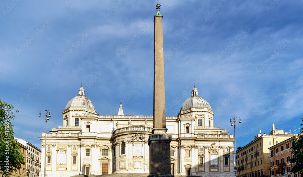 Obelisk of the Plaza called the Esquiline and facade of the papal Basilica called Santa Maria Maggiore, without people in sight and with a blue sky with very light clouds. In Rome, Italy