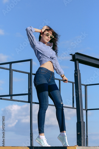 A beautiful girl with sunglasses and torn jeans and a striped shirt. Against the background of large steel structures. Artistic photography. beauty and fashion, style