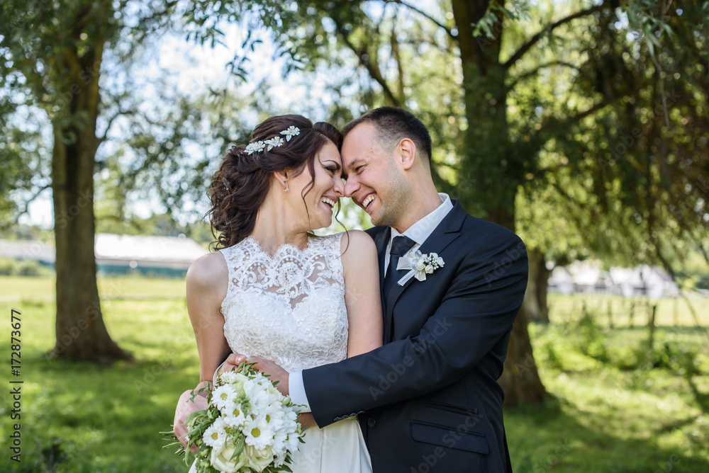 kissing couple in wedding attire with a bouquet of flowers and greenery is in the hands against the backdrop of trees, the bride and groom kissing couple in wedding attire with a bouquet of flowers and greenery is in the hands against the backdrop of trees, the bride and groom