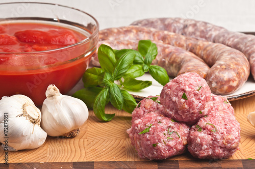 Bamboo cutting board with preparing a homemade pizza with basil, clear glass bowl of crushed tomatoes, garlic heads, hot and sweet italian sausage, organic meatballs, plate and a sprig of basel